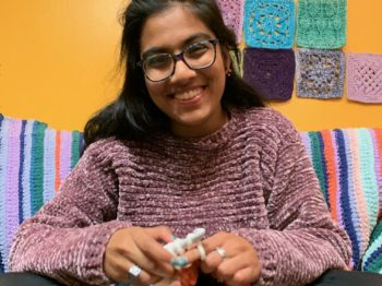 Rima smiles at the camera while crocheting with white and orange yarn.