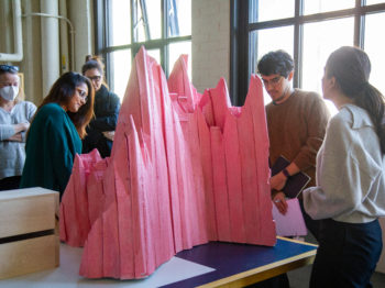 Students stand in a bright warehouse around a pink foam sculpture of a mountain.
