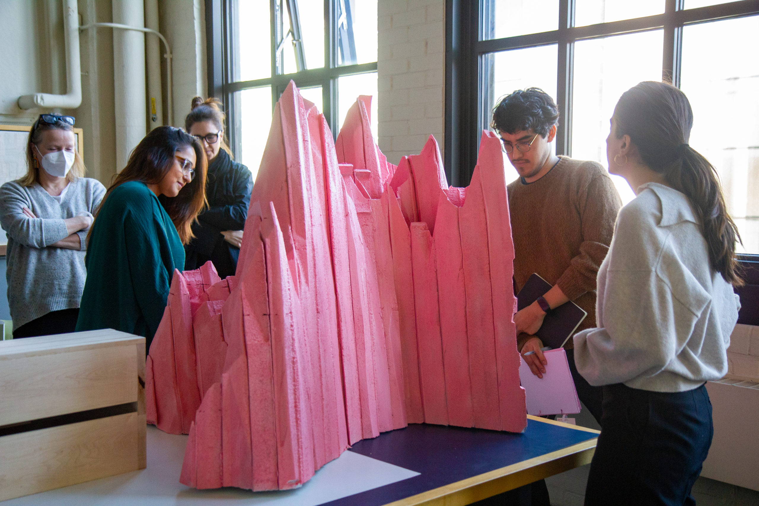 Students stand in a bright warehouse around a pink foam sculpture of a mountain.