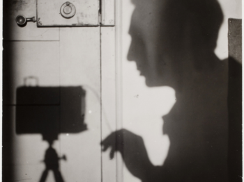 Black and white photograph of the shadow of the photographer cast against a white wood door while pressing the shutter release on their camera.
