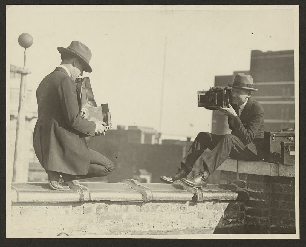 Two photographers taking each other’s pictures with hand-held cameras while perched on a roof, between 1909 and 1932.