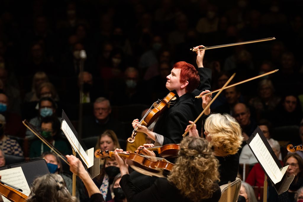 A violinist with bright red hair performs on stage with a chamber orchestra.