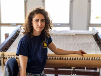 Chloe Bensahel sits facing the camera with one arm resting on a tapestry loom.