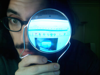 A man holds a mirror up to his face that shows a distorted image of himself on a computer screen.