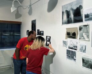 A man and a woman in red t-shirts stand leaning in towards a white with black and white photographs hanging on it as the woman points at a photograph.