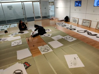 Two participants sit on mats on the floor of the gallery space. Surrounding the participants are large, white pieces of paper covered in graphite, abstract drawings.