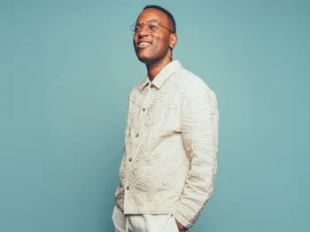 In the midst of a teal background, Michael Mayo smiles while standing casually with his gaze to the horizon.