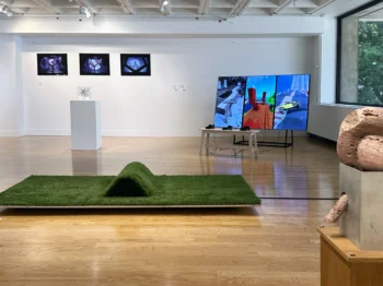 Multiple pieces of artwork including sculptures on pedestals, digital screens, and a low flat astroturf sculpture are installed in a gallery.