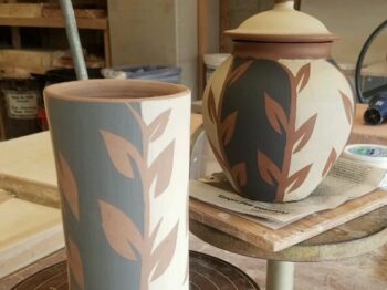 Two handbuilt ceramic pieces on pedestals in a pottery studio. Both have vine and leaf surface decoration.