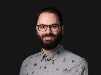 Man with glasses and a beard looks straight to the audience with a faint smile.