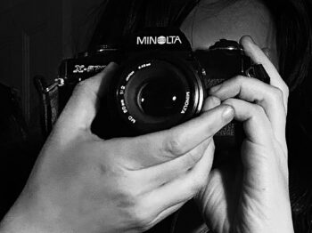 Person holds Minolta film camera facing the viewer; the artist face is obscured by the camera. The two hands holding the camera are featured prominently in the foreground of this black and white photograh.