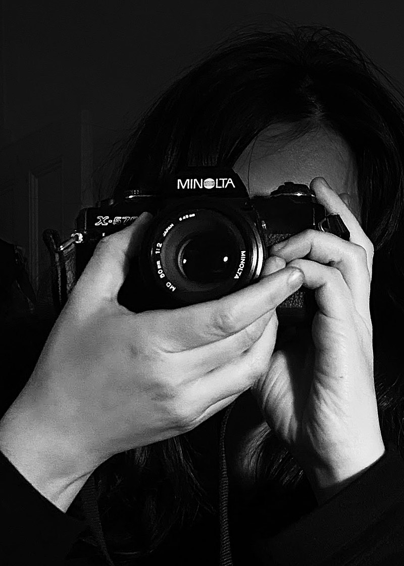 Person holds Minolta film camera facing the viewer; the artist face is obscured by the camera. The two hands holding the camera are featured prominently in the foreground of this black and white photograh.