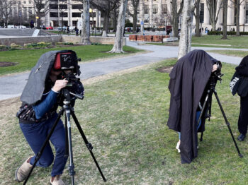 Color photograph of two students standing outside in a public area underneath dark sheets while looking through cameras. Trees and MIT buildings are in the background.