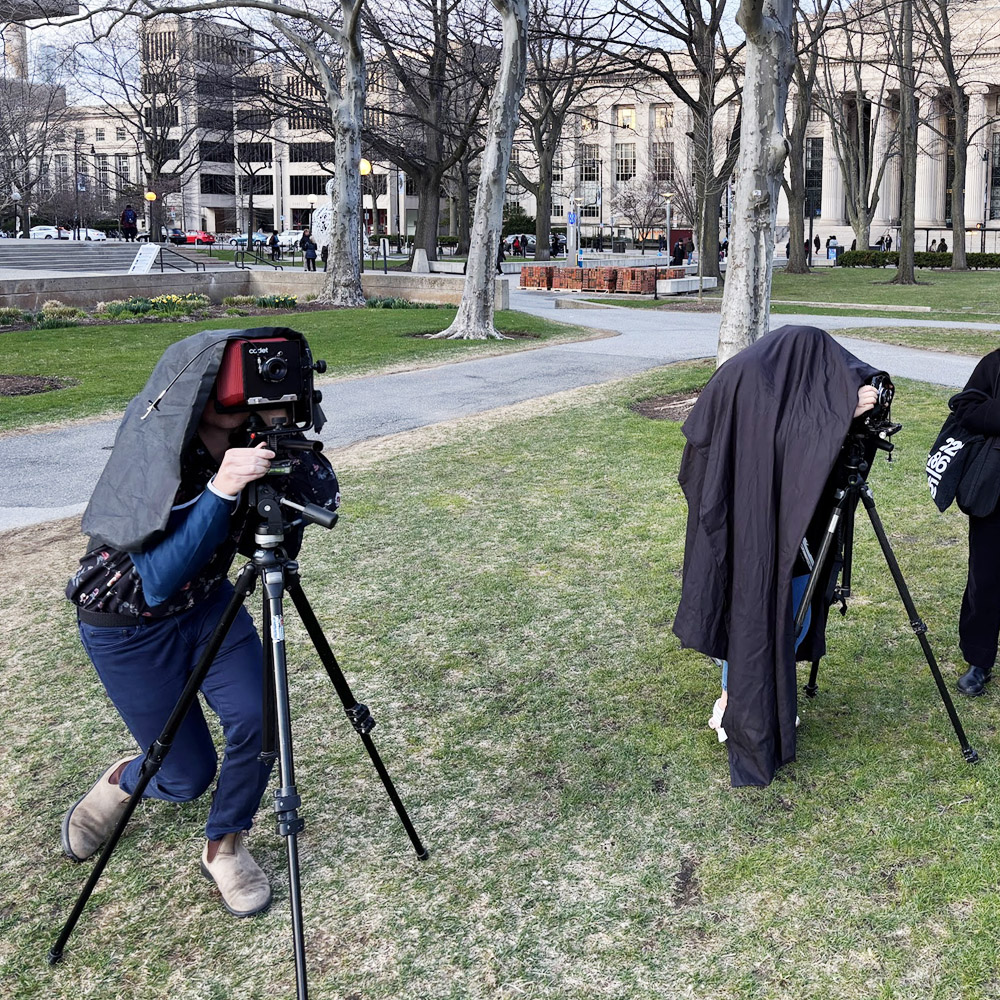 Color photograph of two students standing outside in a public area underneath dark sheets while looking through cameras. Trees and MIT buildings are in the background.