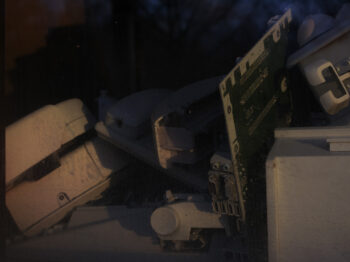Color photograph of different types of photography and computing equipment arranged in a still life manner. The image is dark, almost to the point of the items being abstracted and obscured.