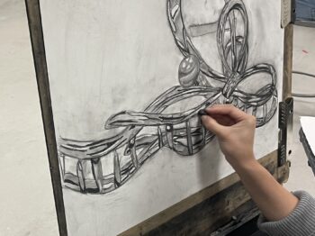 A hand draws with a piece of charcoal on a piece of drawing paper clipped to a drawing board
