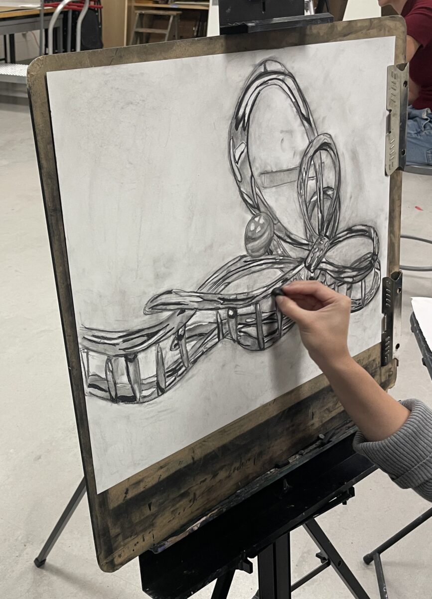 A hand draws with a piece of charcoal on a piece of drawing paper clipped to a drawing board