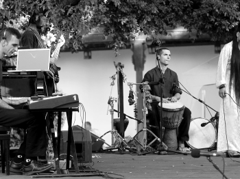 Musicians perform keyboard, drums, and singing on an outdoor stage.