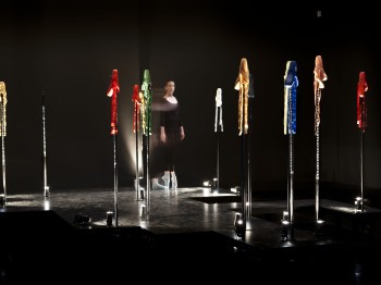 A woman performs on a stage surrounded by colored pointe shoes on poles.