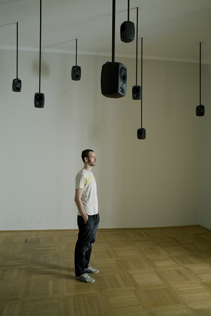 A man stands in a gallery under speakers suspended from the ceiling.
