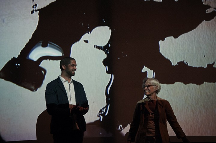 A man and a woman stand in front of a projected image of an ink spill.