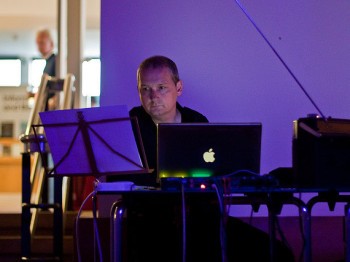 A man sits behind a laptop and a music stand.