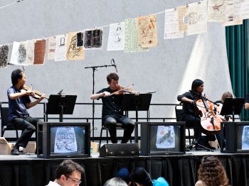 A string quartet performs on a stage with TV screens and hanging artistic prints.