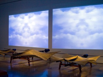 Three wooden sculptures in front of a wall with projections of clouds.
